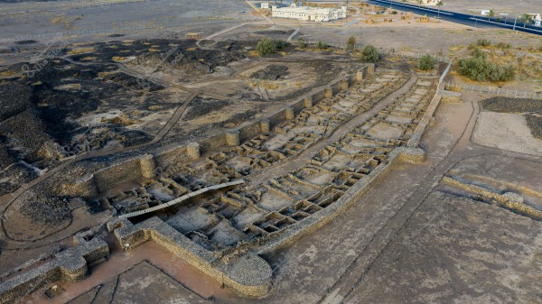 An aerial view of archaeological ruins in a desert area, showing remnants of stone walls arranged in a rectangular shape with clear pathways. The area is surrounded by barren land with some scattered vegetation. Modern buildings and roads are visible in the background.