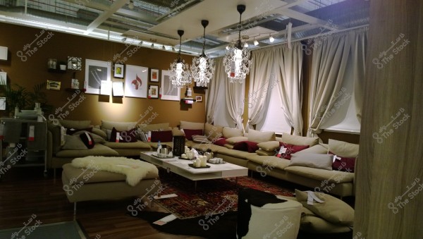 The image shows a modern living room with an elegant and warm design. The room features a large beige corner sofa adorned with a collection of inviting cushions. In the center is a white coffee table decorated with a set of candles and tea utensils. The ceiling features hanging crystal chandeliers, and the walls are adorned with framed artwork. Beige curtains decorate the windows, while a traditional patterned rug covers the wooden floor.