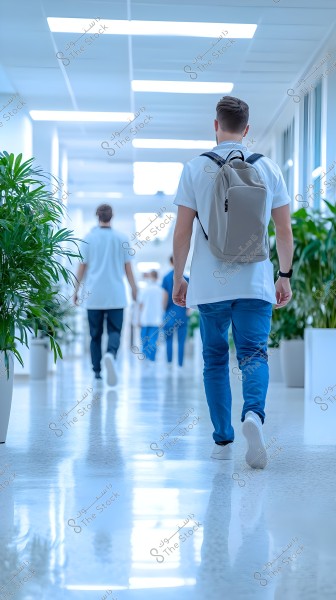 An image of people walking down a well-lit corridor with plants on both sides. The person closest to the camera is wearing a white shirt, blue jeans, and carrying a backpack. Bright lights hang from the ceiling, and there are glass walls that give the corridor a modern and bright appearance.