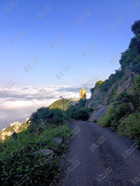 A narrow mountain road with green slopes on either side, leading to buildings perched atop the mountain. The horizon is filled with thick clouds below the road level, and the sky is clear blue above. The lush greenery and density convey a sense of tranquility and natural beauty.