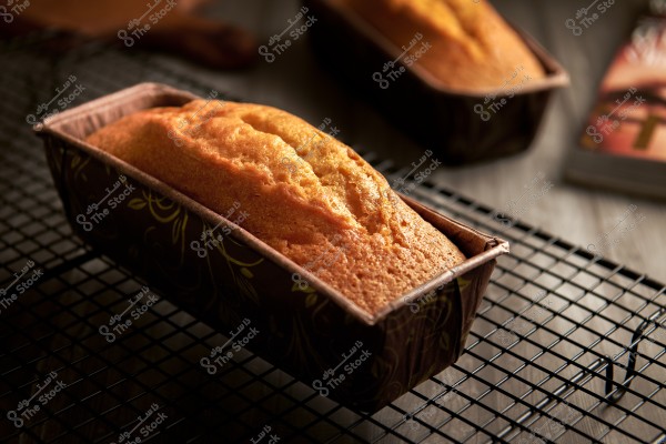 A golden-brown loaf cake in a rectangular pan on a black cooling rack. Another cake is visible in the background, along with a few books, placed on a wooden surface.