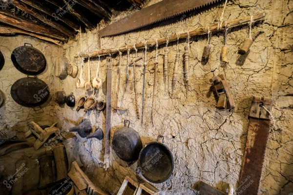 An image of an interior mud wall decorated with vintage farming tools. The wall features hanging saws, metal cooking pots, wooden ladles, and various other wooden and rope tools. The wall displays signs of wear and cracking, reflecting a traditional and rustic atmosphere.