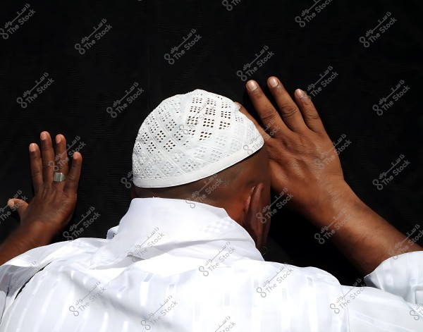 A man placing his hands on Kaaba, wearing a white cap.
