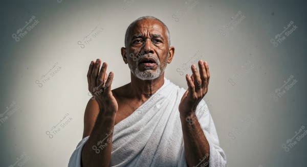 Portrait of an elderly man wearing a white Ihram cloth, raising his hands in a prayer position. His facial expression is serious and focused, suggesting he is performing Hajj or Umrah rituals. The background is neutral, emphasizing the man and his attire.