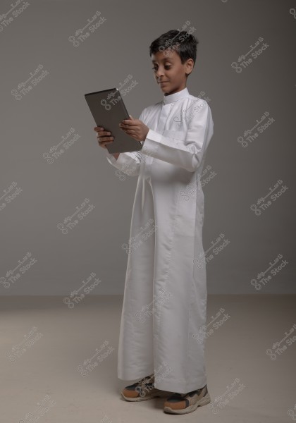 An image of a boy wearing a traditional white thobe, suggesting a Gulf region origin, focusing on a tablet in his hands. He is in an indoor setting with a simple gray background and appears to be slightly smiling.
