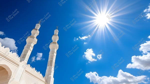 The image features two mosque minarets made of white marble, set against a clear blue sky with a few scattered clouds. The sun shines brightly from the top right corner, casting a radiant glow on the minarets. The architectural ornaments and aesthetic details of the mosque are distinctly visible.
