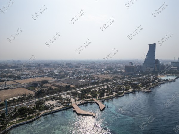 Aerial view of an urban waterfront with blue sea waters and a beachfront promenade. A notable architectural building with a modern design is seen alongside the sprawling city in the background under a clear sky.