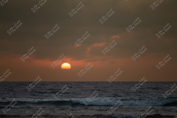 Image of a sunset over the ocean. The bright orange sun is nearing the horizon, with its rays reflecting on the water\'s surface. Clouds cover the sky, adding a dramatic effect to the natural scene.