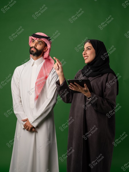 رجل سعودي يرتدي الثوب التقليدي يقف بجانب امرأة سعودية ترتدي العباءة, تحمل جهازًا لوحيًا وتشير بإصبعها, والخلفية خضراء.