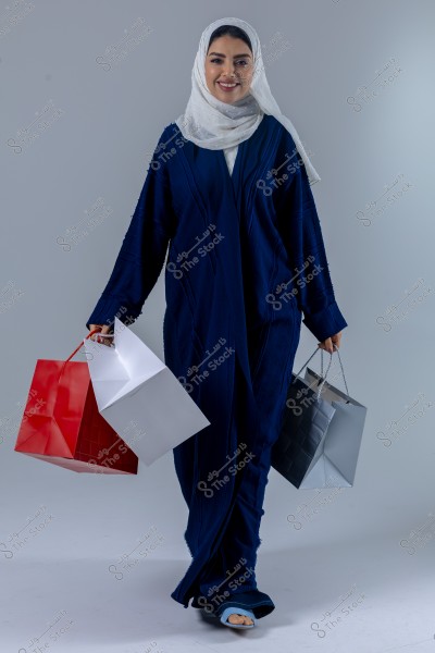Image of a woman wearing a dark blue abaya and a white hijab, carrying shopping bags in various colors including red, white, and gray. She is smiling and standing against a neutral gray background. The attire suggests a traditional look typical of women in some Gulf countries.