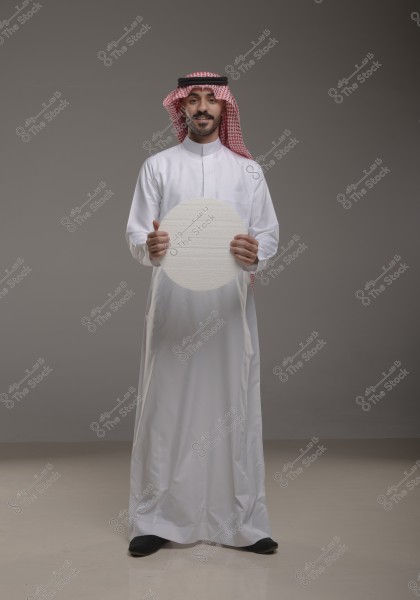 The image shows a man wearing a traditional white thobe and a red and white checkered ghutra with a black agal. He is standing in a studio with a grey background, holding a round white board with his hands.