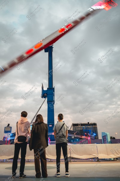 Three people stand in front of a towering amusement ride in an entertainment park. The sky is cloudy in the background, and a blue crane supports a fast-spinning platform, with colorful lights illuminating the scene.
