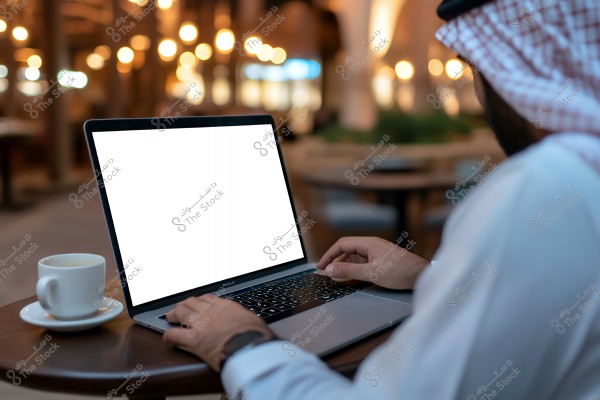 A man wearing a white ghutra and agal is sitting in an outdoor café-like setting using a laptop with a blank screen. On the table, there\'s a white coffee cup. The background is warmly lit and blurred with lights.