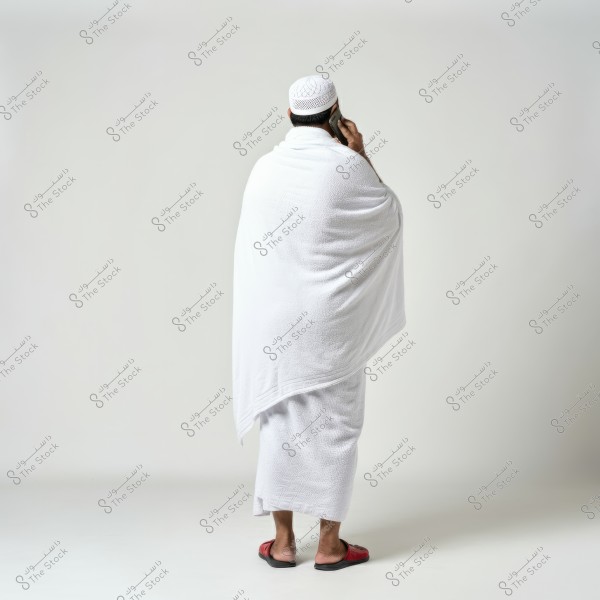 The image shows a man wearing a white Ihram, standing with his back to the camera. He is wearing a white head cover and using a mobile phone. He is wearing red sandals and standing against a simple white background. The clothing indicates that the image may be related to the rituals of Hajj or Umrah in Saudi Arabia.