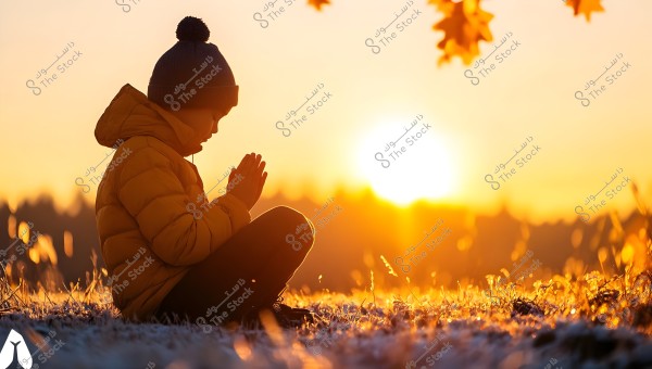 A child wearing a yellow winter jacket and a knitted hat sits in a frost-covered field at sunrise. The child holds their hands together in front of their face, appearing to be praying or meditating. The warm colors of the sun fill the background, creating a serene and beautiful scene.