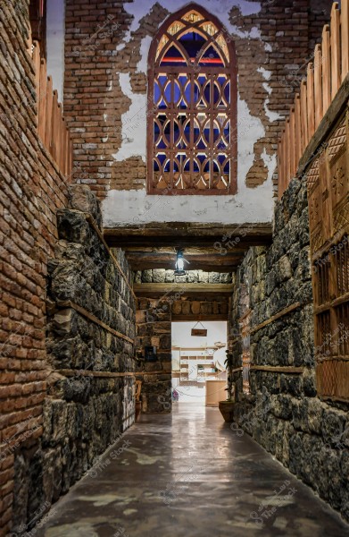 A narrow interior corridor in a traditional building with architectural elements of stone and brick. The inner walls are a combination of brick and dark stone. An ornate wooden design is visible through the upper window of the corridor, and the entrance at the end leads to a well-lit room with display shelves.
