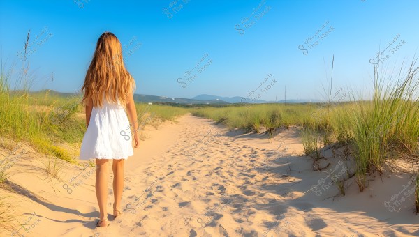 The image features a girl walking barefoot on a sandy path surrounded by tall green grass. She is wearing a short white dress, and her long brown hair cascades down her back. The sky is clear and blue, with low hills visible in the background.