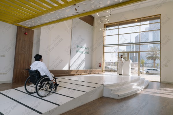 A person "with disabilities" in a wheelchair moves towards an elevated podium in a modern lecture hall. There is a person standing on the podium with large windows in the background revealing city views.