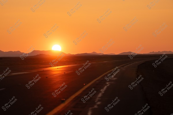 A sunset scene over a desert road with the horizon aglow in orange hues as the sun sets behind distant mountains. The road stretches towards the horizon, creating a sense of distance and tranquility.