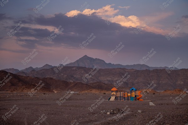 A landscape view of a desert mountainous area at sunset, featuring a children\'s playground in the foreground with slides and play equipment in orange and blue colors. The mountains are in the background under a partially cloudy sky, tinted with sunset hues.