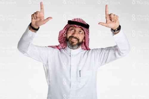 بورتريه رجل سعودي يقف على خلفية بيضاء، يرتدي الثوب، يقف امام الكاميرا ويقوم بعمل حركات بيديه، يمد يديه الى اعلى و ويشرح بأصابعه ضمن اطار محدد بحماس