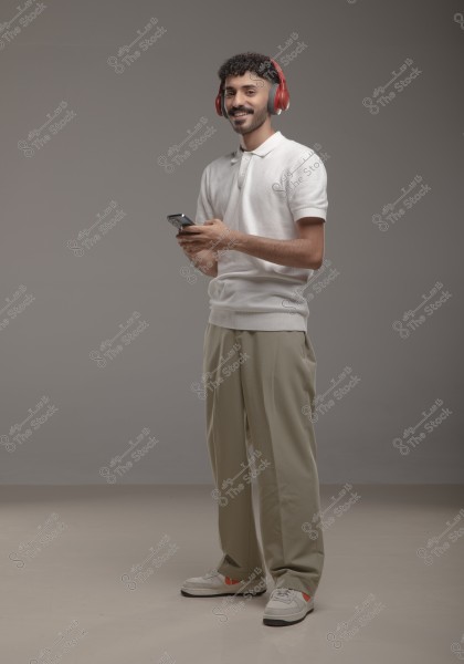 A man is standing in a studio wearing red headphones, a white polo shirt, and beige pants. He is smiling and holding a smartphone. The floor is grey, and the walls are a light grey color, giving the image a modern and clean feel.