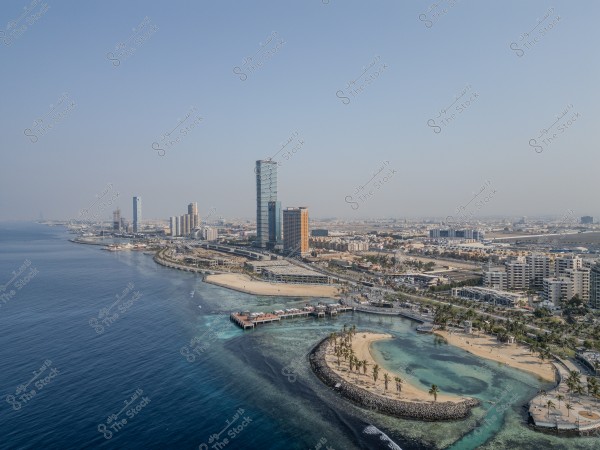 An aerial view of a modern waterfront in a city located on the Red Sea coast, featuring a cluster of tall buildings and hotels. Palm trees are lined along the sandy beach, with a stretch of roads and tourist facilities. The clear blue waters extend to the horizon under a clear sky.