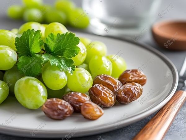 A plate containing fresh green grapes and several dark brown dates. Some fresh green leaves are in the center of the plate. The plate is placed on a gray table with cutlery beside it.
