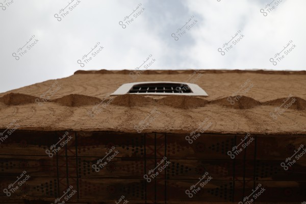 The image shows an angle of a traditional mud-brick building with a window featuring iron bars and decorative traditional patterns at the base. The sky is cloudy. The design reflects the traditional architectural style of Gulf countries, such as Saudi Arabia.