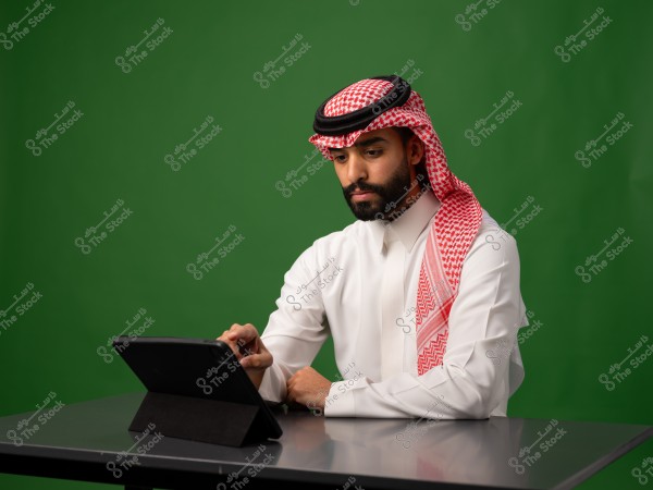 رجل يرتدي الزي السعودي التقليدي يستخدم جهاز تابلت على طاولة, وخلفه خلفية خضراء.