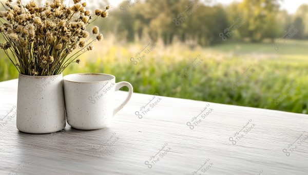 A cup and a vase containing small dried flowers placed on a white wooden table, with a blurred nature background filled with trees and plants.
