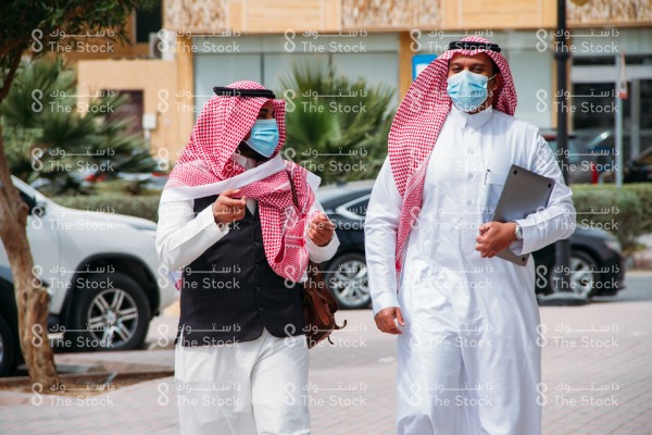 رجلان سعوديان يرتديان الزي السعودي التقليدي و الكمامة يمشيان على الرصيف بالشارع نهارًا يتناقشان في طريقهما للعمل بينما يحمل أحدهم شنطة ظهر والآخر يمسك بالابتوب وتظهر من خلفهم المباني والسيارات والأشجار