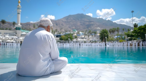 A man wearing a white robe and a head covering sits by the edge of a large pool of water, with a mountainous backdrop and a mosque featuring a minaret. The scene is sunny, with people in white clothing standing on the other side of the pool, and palm trees and greenery in the background.