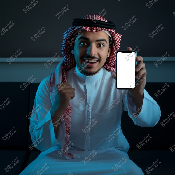 An image of a man sitting on a sofa in a dimly lit room, wearing traditional Gulf attire, including a white thobe, red and white ghutra, and a black agal. The man is smiling and holding a smartphone with a blank white screen.
