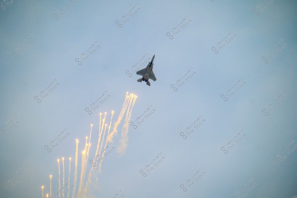 A fighter jet flying in the sky, releasing multiple bright flares downward. The sky is clear blue, and the flares trail behind the aircraft in bright streaks.