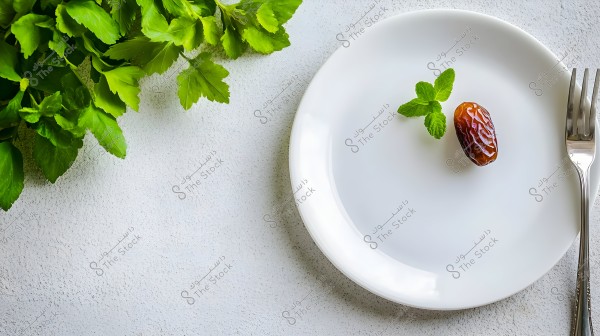 An image of a white plate with a single date and a green mint leaf, placed on a white surface. A silver fork is visible beside the plate. In the top left corner, there are some green herbs.