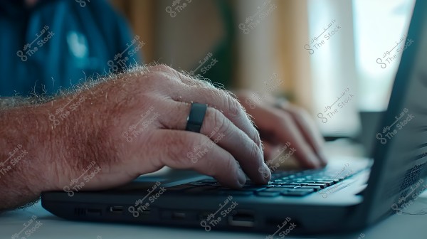 The image shows a man\'s hands with a black ring on one finger, typing on a laptop keyboard. The focus is on the hands and keyboard, with a blurred background suggesting a bright window and a dark-colored shirt.
