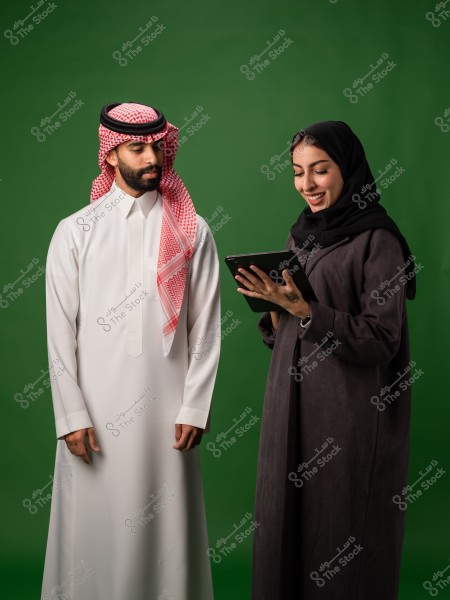 رجل يرتدي الزي الخليجي التقليدي يقف بجانب امرأة محجبة تمسك بجهاز لوحي على خلفية خضراء.