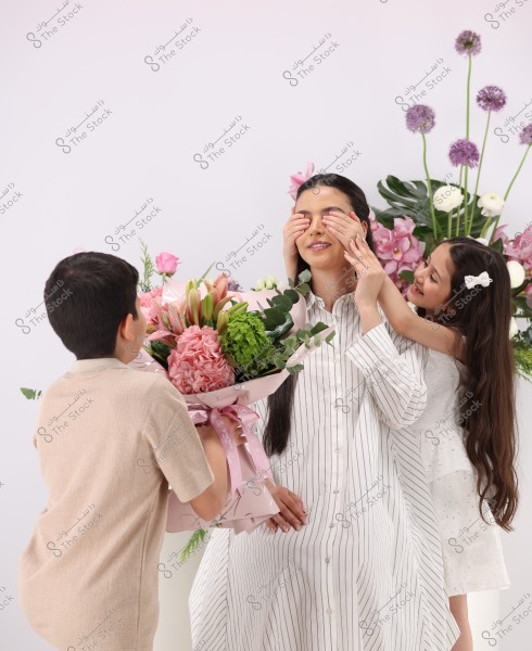 An image of a seated woman wearing a white and black striped dress surrounded by flowers. A boy and a girl are presenting her with a large bouquet and covering her eyes with their hands. Behind them, there is a beautiful arrangement of colorful flowers against a white background.