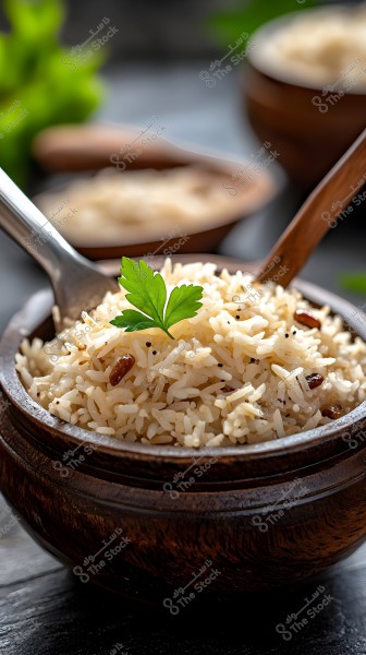 A bowl of cooked white rice in a wooden bowl, garnished with a small parsley leaf on top. The rice contains small pieces of spices and dark pieces, possibly raisins. The wooden bowl is placed on a gray table surface, with another bowl of rice slightly visible in the background.