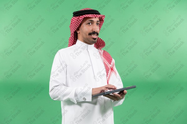 رجل "من ذوي الاعاقة" يرتدي الثوب السعودي والشماغ الأحمر يستخدم حاسوبًا لوحيًا بخلفية خضراء.