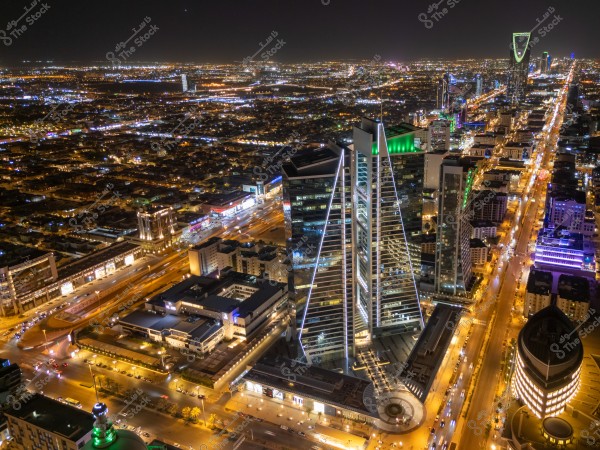 Aerial night view of Riyadh, Saudi Arabia, showcasing the city skyline illuminated with the lights of buildings and bustling streets. The image prominently features a variety of modern skyscrapers, including the iconic Kingdom Tower, surrounded by brightly lit buildings. The city stretches into the horizon with sparkling city lights visible throughout.