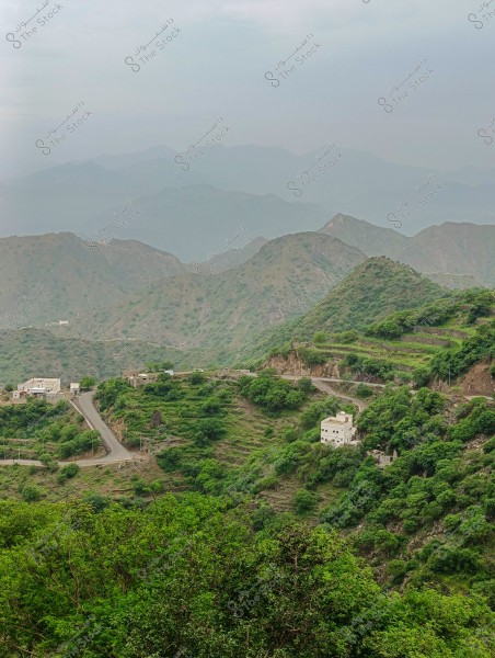 A scenic view of sprawling green mountains with winding roads and scattered buildings, with foggy mountains in the background under a cloudy sky.