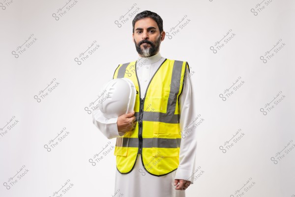 A portrait of a man wearing a white traditional garment and a bright yellow safety vest with gray reflective stripes. He is holding a white construction helmet in his right hand. The background is white, highlighting the figure in the image. The design of the garment suggests it might be from the Arabian Peninsula.