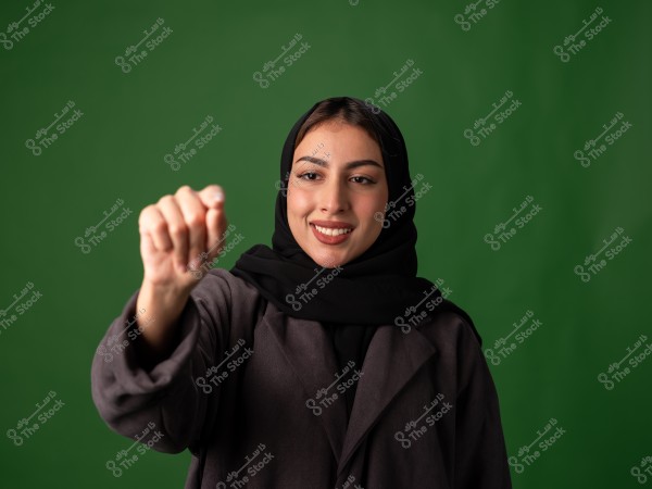 The image shows a smiling woman wearing a black headscarf and a dark-colored abaya. She is standing in front of a green background while making a gesture with her right hand. Her facial expression suggests confidence and positivity.