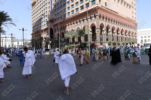 The image shows a group of people wearing white Ihram clothing gathered in a square in Mecca, Saudi Arabia. Next to them is a large building with distinctive Islamic architectural design and rows of palm trees. The sky is clear, and the atmosphere is sunny.