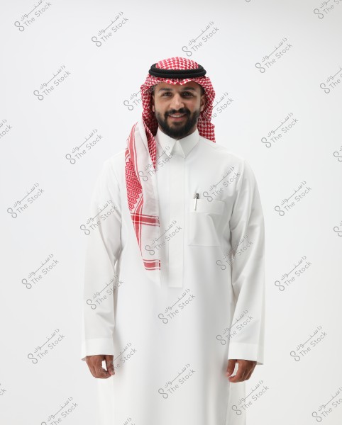 A portrait of a smiling man standing, wearing traditional Saudi attire including a white thobe and a red and white ghutra with a black igal.