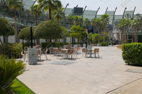 An open outdoor plaza featuring wooden tables and chairs, surrounded by trees and greenery. The scene includes palm trees and orchard-like trees with rounded foliage, set against modern architecture in the background, suggesting a comfortable setting for relaxation or outdoor dining.