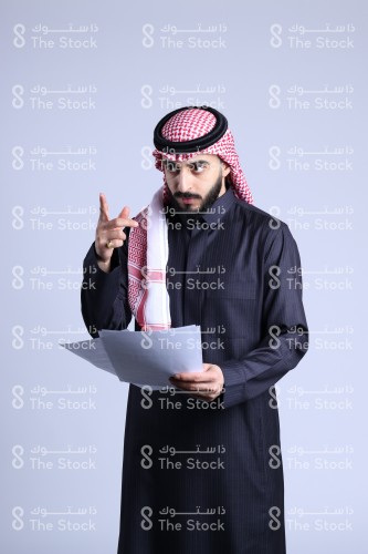 Portrait of a Saudi man in traditional Saudi uniform holding a set of papers while pointing his other hand