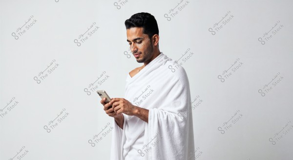 A photo of a man wearing a white wrap using a smartphone. The man is looking attentively at his phone and is dressed in traditional white clothing that may be associated with Ihram, suggesting a religious context like Hajj or Umrah. The background is plain white.